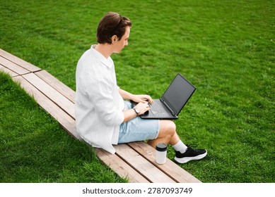 Freelancer doing remote work on a laptop outdoors. Man sitting on a wooden bench next to green grass, typing online with a coffee cup nearby. High quality photo