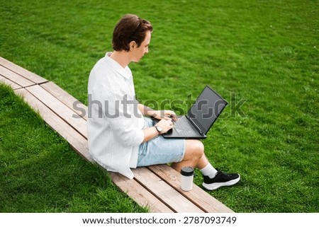 Freelancer doing remote work on a laptop outdoors. Man sitting on a wooden bench next to green grass, typing online with a coffee cup nearby. High quality photo
