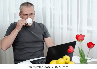 Freelancer drinking coffee while using laptop.Middle aged man with spectacles sipping coffee from a white cup while working on a black laptop at home at a round table with fresh flowers.