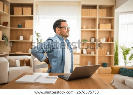 Freelancer feeling backache after long hours of working on a laptop and sitting on a chair in his home office, he is touching his back with hands and suffering from back pain