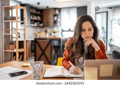 Freelancer focused on taking notes in a notebook while working from home, using a laptop to manage online business tasks in a cozy home office setting