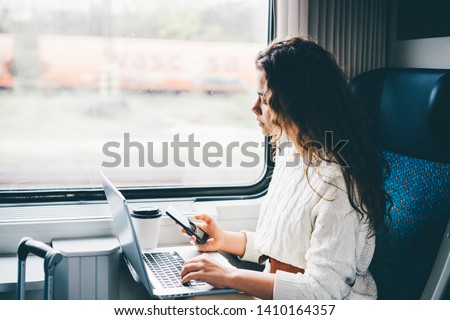 Freelancer girl working with laptop in the train. Girl looking to the phone in her hand. Business travel or technology concept.