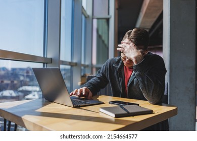 A freelancer in glasses looks into a laptop and he is angry while sitting at a table with a laptop and a notepad in a cafe during the day. Unlucky day. Working remotely from the office.
