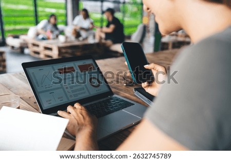 Freelancer holding smartphone while working on laptop with business dashboard, people networking in background, relaxed atmosphere in shared tropical coworking space, productivity on the go