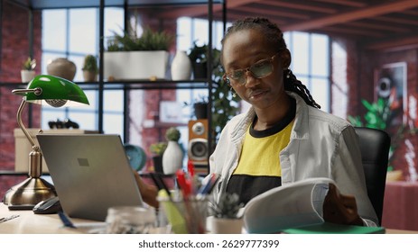 Freelancer at home checking analytics statistical data sets on laptop, comparing with paperwork files. Woman optimizing key performance indicators on notebook by crosschecking with documents, camera B
