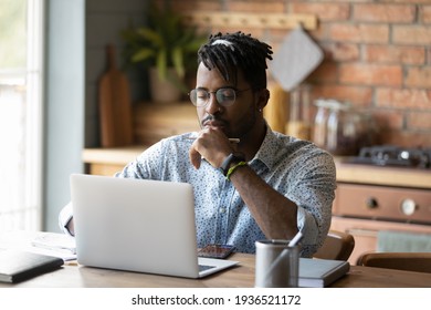 Freelancer At A Job. Young African Man Hipster Think By Laptop At Home Office Prepare Report Electronic Document Use Open Source Web App. Focused Black Male Student Read Web Search Result On Pc Screen