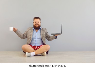 Freelancer Online Distance Work Concept. Funny Fat Bearded Man In A Jacket And Red Shorts With A Laptop On A Gray Background.