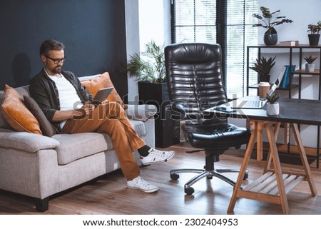 Freelancer sitting on sofa, looking relaxed as he uses tablet PC. Laptop on desk and empty chair can be seen nearby in room. Concept of relaxation after work, and ease and comfort of working from home
