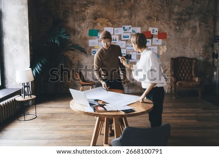 Freelancer team analyzes project data on printed charts, discussing strategic insights and digital workflow around round desk in loft office, showing collaboration and creative process.