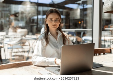 Freelancer typing on laptop in cozy café. Modern workspace with coffee, phone, and stylish accessories. Productive remote work scene. Woman working remotely in a café, using laptop with phone