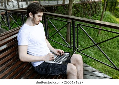 Freelancer using computer outdoors in city park. White t-shirt, black shorts, relaxed setting.