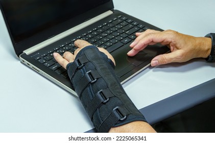 Freelancer Woman With Broken Wrist Is Typing On Laptop On Keyboard. Woman's Left Hand Is In Black Orthosis Instead Of Cast. Hands On Laptop Keyboard Close Up