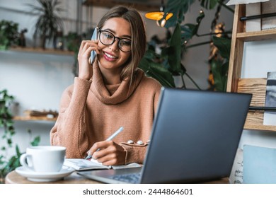 Freelancer woman sitting in a coffee shop with a laptop on the table while making notes and talking on cellphone. Focused businesswoman in casual outfit drink coffee while working remotely in cafe - Powered by Shutterstock