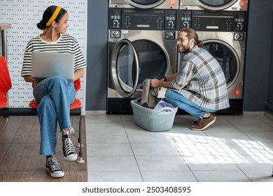 Freelancer woman using laptop while man doing laundry at laundromat - Powered by Shutterstock