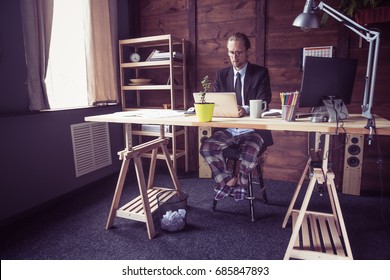 Freelancer At Working Place At Home. Man In Jacket Working With Laptop, His Legs In Pajamas Under Table. Toned Image.