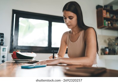 Freelancer writes in notebook at kitchen table with watermelon and phone nearby, expressing balance between tech, health and focused analog productivity.