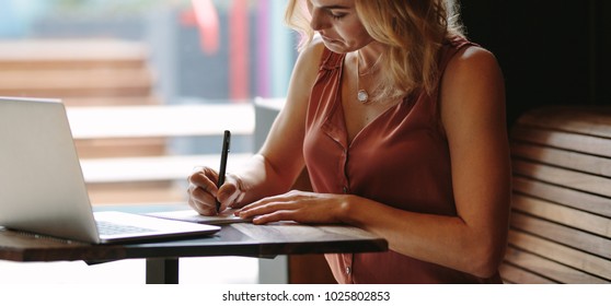Freelancer writing notes sitting at a coffee table. Woman working on her laptop computer at a coffee shop.