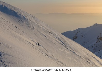 A freerider snowboarder descends a wide slope against the background of mountain ranges and the sunset, active recreation in the backcountry