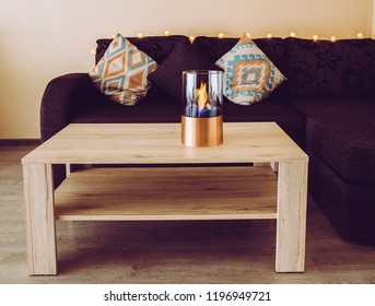 Freestanding Portable Glass And Copper Metal Fireplace Burning Bio Ethanol Gas On Living Room Brown Natural Wooden Table, Black Coach On The Background. Autumn Comfortable Colorful Cozy Home Set.
