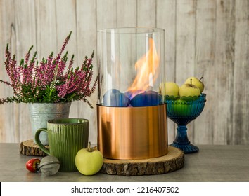 Freestanding Portable Glass And Copper Metal Fireplace Burning Bio Ethanol Gas On Wooden Table With Tee Cup, Blue Bowl With Green Apples. Autumn Set Comfortable Colorful Warm Cozy Home Concept.