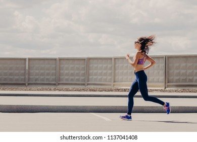 Freeze Action Of Beautiful Slim Woman Has Morning Jogging Exercise, Wears Top And Leggings, Breathes Fresh Air While Runs, Enjoys Sport, Has Healthy Lifestyle. Female Runner Has Workout Outdoor