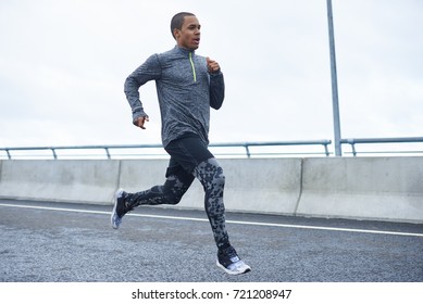 Freeze action shot of confident young black athletic man wearing trendy running shoes and sports clothes exercising outdoors. Picture of dark-skinned sportsman running on autumn day at stadium