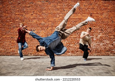 Freeze frame of male breakdance performer doing handstand pose with team against brick wall outdoors