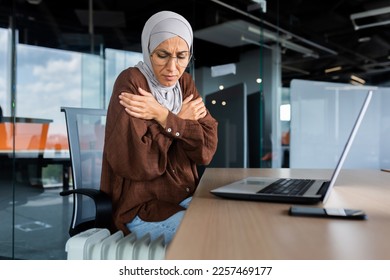 Freezing arab woman inside office, business woman in hijab cold indoor heating and heating not working, using laptop in work at workplace, sitting trying to warm. - Shutterstock ID 2257469177