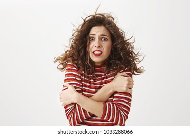 It is freezing cold outside. Indoor shot of poor attractive girl with curly hair that flows on wind, hugging herself and trembling, standing without coat on windy weather over gray background