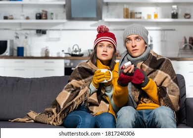 freezing couple in warm hats, wrapped in plaid blanket, watching tv while sitting in cold kitchen - Powered by Shutterstock