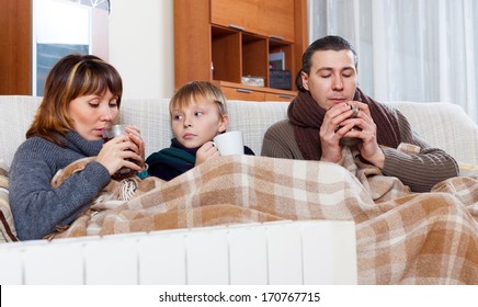 Freezing Family Of Three With Cups Of Tea Warming Near Warm Radiator In Home