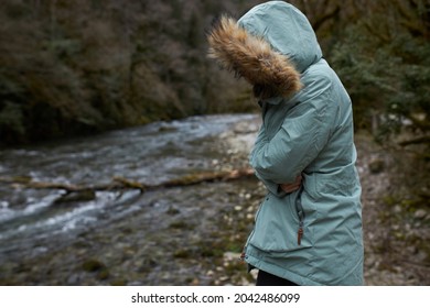 Freezing girl wrapped up in blue parka coat with face hidden under furry hood, hugging herself to keep warm inside, standing against narrow mountain river with fast flow, forest growing on its banks
