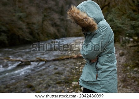 Freezing girl wrapped up in blue parka coat with face hidden under furry hood, hugging herself to keep warm inside, standing against narrow mountain river with fast flow, forest growing on its banks
