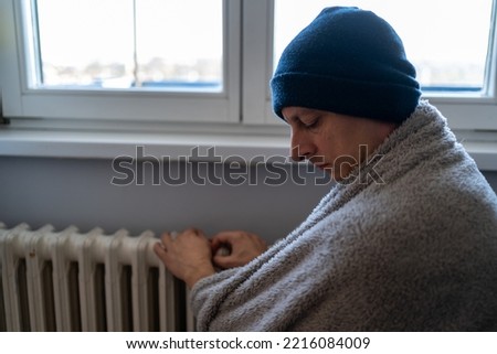 freezing man in warm hats and gloves looking at camera while sitting on sofa in cold living room. Middle Aged Man Trying To Keep Warm Under Blanket At Home. Heating season. Cold adult man energy