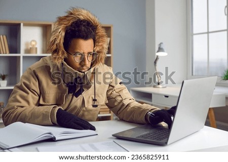 Freezing man in warm jacket and gloves working at computer in office. Unhappy warmly dressed businessman freezing in office since heating problems or power crisis at cold season