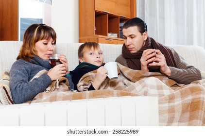 Freezing Parents And Teenage Son With Cups Of Tea Warming Near Warm Calorifer In Home