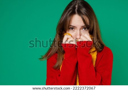 Freezing young brunette woman 20s wearing basic casual knitted red sweater covering face with yellow scarf trying to keep warm looking camera isolated on bright green color background studio portrait