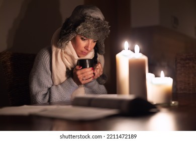 Freezing Young Woman In Winter Clothes Warms Her Hands On Cup Of Tea And Lights With Candles As Energy Blackouts Cause Electricity Outages.