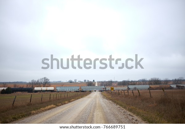 Freight train\
crossing a straight rural dirt road at a level crossing in a\
receding view perspective under grey\
cloud