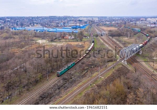 A freight train with a green electric
locomotive rides along the railroad tracks.  View from the height
of the drone.  Overcast.