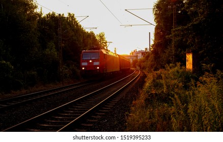 Freight Train With Modern Electric Locomotive On A Curved Main Line Railway Track In Iserlohn-Letmathe Sauerland Germany. Sunset Atmosphere With Warm Evening Light, Overhead Lines, Weeds And Trees.