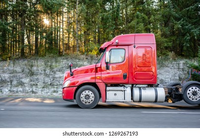 Freight transport by semi trucks in America - the main type of logistics. Red big rig semi truck with cargo on flat bed semi trailers running on highway with winter frosty trees with sun