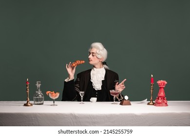 French breakfast. Portrait of young elegant man in peruke and vintage jacket sitting at table isolated on dark green background. Retro style, comparison of eras concept.