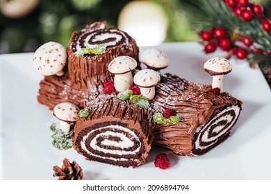 French dessert called Yule log or bûche de Noël with merengue mushrooms and mint leaves on top of chocolate glazing. Placed in front of Christmas tree. Decorated for Christmas Holidays or New year - Shutterstock ID 2096894794