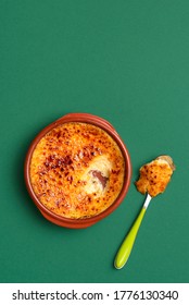 French Crème Brûlée Dessert In A Clay Tray With Spoon, On Green Background. Top View Of Burnt Cream With Caramel Crust. Custard With Burnt Sugar.