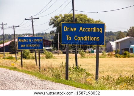 French, English Information road Signs, Drive According To Conditions. On Canadian rural country roadside with farms in the background, Ontario Canada
