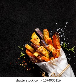 French fries,  baked fries from different types and colors of potatoes sprinkled with herbs and spices in paper bag on a black background, top view, copy space