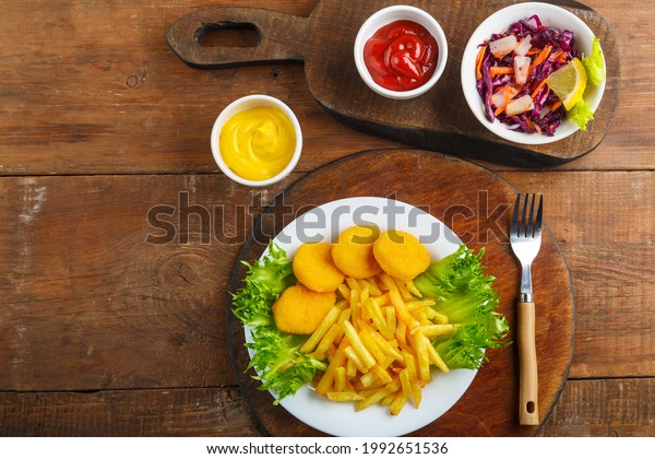French fries with chicken nuggets next to cheese\
sauce and ketchup in a gravy boat and salad on wooden planks next\
to a fork. Horizontal\
photo