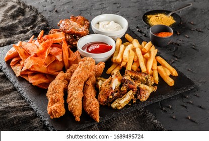 Pommes frites, Kartoffeln, gegrilltes Fleisch und Fisch, Hühnernuggets, scharfe Kartoffelchips, Ketschup und Mayonnaise auf schwarzem Hintergrund. Fast-Food-Snacks, Nahaufnahme
