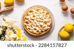 French lemon tart with churned meringue on marble table with some lemons, eggs, and spring yellow flowers in the corner, top view, horizontal image